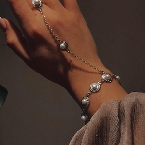 May include: A silver hand chain bracelet featuring a ring and bracelet connected by a delicate chain. The jewelry is adorned with small, round pearls set in ornate, circular frames. The design is elegant and classic.