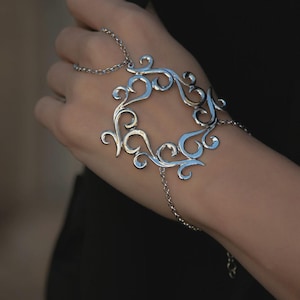 May include: Silver-toned hand chain bracelet featuring an openwork design with swirling, symmetrical patterns. The bracelet connects to a ring and wrist chain. The jewelry is worn on a hand against a dark background.