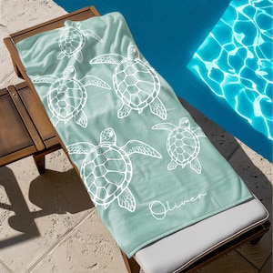 May include: A light teal beach towel with a white turtle pattern, resting on a wooden lounge chair next to a pool. The towel features a repeating design of sea turtles and the name "Oliver" in cursive.