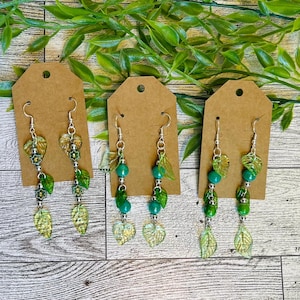 May include: Three pairs of dangle earrings on brown tags. Each earring features silver hooks, green beads, and leaf-shaped charms in shades of green and gold. The earrings are displayed on a wooden surface with green foliage.