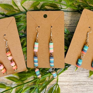 May include: Three pairs of colorful beaded earrings displayed on brown earring cards. The earrings feature a variety of pastel colors, including pink, orange, blue, and green, with gold-colored accents. The earrings are hanging on silver-colored hooks.