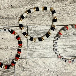 May include: Three beaded bracelets with different color schemes and text. One bracelet spells "PHANTOM" with black, red, and gold beads. Another has black, white, and gold beads. The third spells "ANGEL OF MUSIC" with red heart beads.