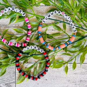 May include: Three handmade bracelets with colourful beads and white letter beads spelling "PINK LADIES", "SUMMER NIGHTS", and "BIRDS". The bracelets feature pink, orange, blue, red, black, and grey beads. The bracelets are displayed on a bed of green leaves.
