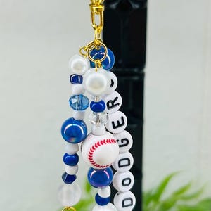 May include: A beaded keychain featuring a gold-tone lobster clasp, blue and white beads, a miniature baseball charm, and white letter beads spelling DODGER. The design includes silver-tone charms shaped like a baseball bat and a glove.