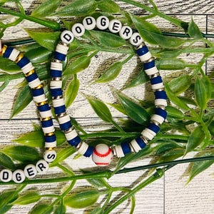 May include: Two beaded bracelets with a baseball theme. One bracelet spells out "DODGERS" in white letter beads. The other bracelet has a baseball bead. The bracelets are blue, white, and gold.