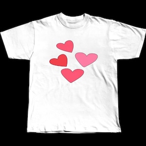 May include: White t-shirt with four hearts in shades of red and pink. The hearts are scattered across the front. The t-shirt has a crew neck and short sleeves. The fabric appears to be cotton.