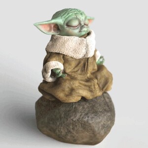 Op de afbeelding: Een groene figuur van een personage met grote oren en gesloten ogen, gekleed in een bruine mantel met een witte kraag. Het beeldje zit op een grijze steen, met een neutrale achtergrond.