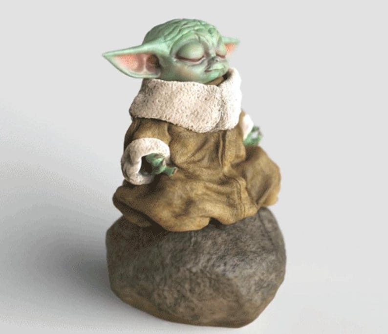 Op de afbeelding: Een groene figuur van een personage met grote oren en gesloten ogen, gekleed in een bruine mantel met een witte kraag. Het beeldje zit op een grijze steen, met een neutrale achtergrond.