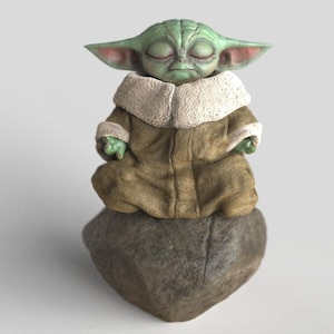 Op de afbeelding: Een groen beeldje van een personage met grote oren, gekleed in een bruine mantel met een witte kraag, zittend in een meditatieve pose op een grijze steen. Het beeldje heeft gesloten ogen en kleine groene handen.