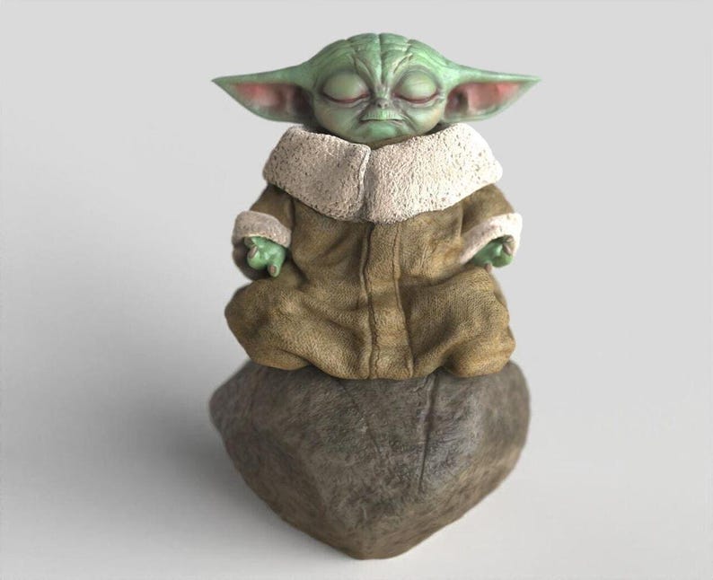 Op de afbeelding: Een groen beeldje van een personage met grote oren, gekleed in een bruine mantel met een witte kraag, zittend in een meditatieve pose op een grijze steen. Het beeldje heeft gesloten ogen en kleine groene handen.