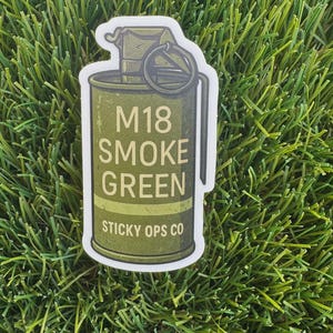 May include: A green sticker featuring a stylized illustration of an M18 smoke grenade. The text "M18 SMOKE GREEN" is printed on the grenade, with "STICKY OPS CO" below. The sticker is set against a background of green grass.