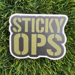 May include: A sticker with the words "STICKY OPS" in a stencil font. The text is olive green on a dark gray background with a white border. The sticker is on a green, grassy surface.