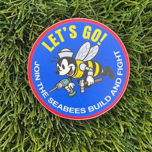 May include: A round sticker with a cartoon bee wearing a sailor hat and yellow and black stripes. The sticker has the text "LET'S GO!" and "JOIN THE SEABEES BUILD AND FIGHT" in yellow and white.