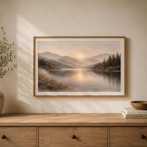 May include: A framed landscape print featuring a serene lake surrounded by pine trees and misty mountains at sunrise. The artwork displays soft golden light reflecting on the water, housed in a light wood frame hanging on a neutral wall.