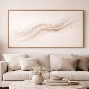 May include: A neutral-toned living room with a large framed artwork featuring flowing, abstract lines in shades of beige and brown. The artwork hangs above a cream-colored sofa with several pillows. A round coffee table and a lamp complete the scene.