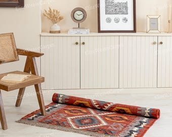 May include: A rolled-up, rectangular rug with a geometric pattern in shades of red, gray, yellow, and blue. The rug is partially unrolled on a light-colored floor, showcasing its design and fringed edges. The rug is a decorative floor covering.