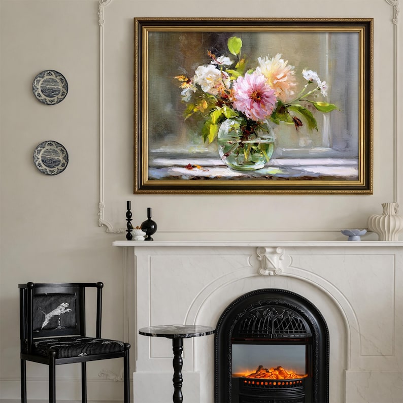 Op de afbeelding: Een ingelijst bloemstilleven met een gouden lijst, met een glazen vaas gevuld met roze, witte en gele bloemen. Het schilderij hangt boven een witte open haard met een zwarte vuurhaard. Decoratieve borden en een zwarte stoel zijn ook zichtbaar.