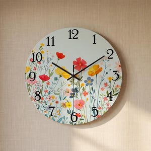 Puede incluir: Un reloj de pared redondo con fondo claro y un estampado botánico colorido de flores silvestres rojas, amarillas y azules. La esfera muestra números arábigos negros del 1 al 12 e incluye finas manecillas negras para horas y minutos.