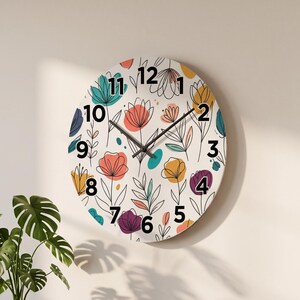 Op de afbeelding: Een ronde wandklok met een witte achtergrond en een kleurrijk bloemenpatroon van blauwgroene, koraalkleurige, gele en paarse bloemen en bladeren. De wijzerplaat toont dikke zwarte cijfers van één tot twaalf en twee dunne zwarte wijzers.