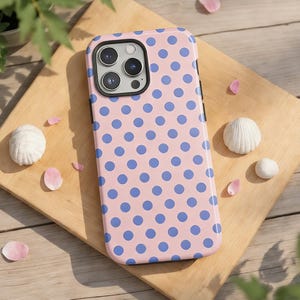 May include: A pink phone case with a blue polka dot pattern. The case is on a wooden cutting board, surrounded by seashells and flower petals. The phone case has a triple-lens camera.