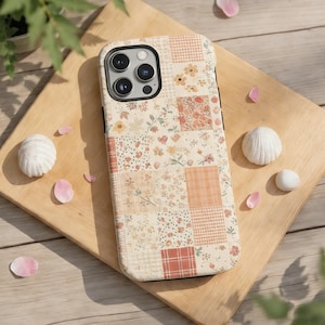Op de afbeelding: Een telefoonhoesje met een patchwork-ontwerp met bloemen- en ruitpatronen in crème-, perzik- en roodtinten. De hoes heeft een zwarte rand en een uitsparing voor de camera. De hoes ligt op een houten snijplank.