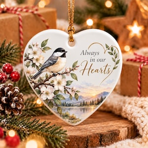May include: A heart-shaped white ceramic ornament hanging from a gold cord. It features a black-capped chickadee perched on a flowering branch, a landscape with mountains and a lake, and the gold text Always in our Hearts.