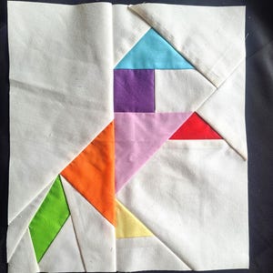 May include: A square fabric patchwork block with a geometric design. The design incorporates white, orange, green, yellow, purple, pink, red, and blue fabric pieces. The block is likely part of a larger quilt or textile project.