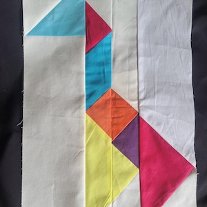 May include: A colorful patchwork quilt block featuring geometric shapes in shades of blue, red, orange, yellow, and purple against a white background. The design includes triangles and diamond shapes, creating a modern abstract pattern. The fabric appears to be cotton.