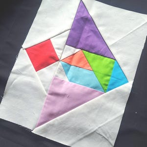 Könnte beinhalten: Ein farbenfroher Patchwork-Quiltblock mit geometrischen Formen. Das Design umfasst ein rotes Quadrat, ein lila Dreieck und verschiedene andere Formen in Orange, Grün und Blau, alles vor einem weißen Hintergrund. Der Block ist aus Stoff.