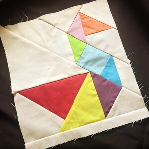 May include: A geometric quilt block featuring a patchwork design of triangles in various colors, including red, yellow, purple, blue, green, pink, and orange. The triangles are set against a white background.