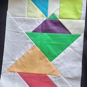 May include: A patchwork quilt block with a geometric design. It features triangles and squares in white, yellow, blue, green, purple, orange, and red. The fabric is likely cotton, and the block is ready for quilting. The block is approximately 30 cm x 30 cm.