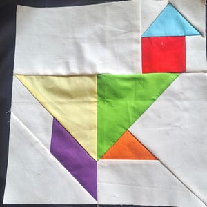 May include: A patchwork quilt block with a geometric design. It features a red square and a blue triangle, resembling a house, alongside yellow, green, orange, purple, and white fabric. The square block is ready for quilting, with measurements in centimetres.
