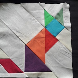 May include: A square quilt block with a geometric design. The block features white fabric sections and colourful accents in red, orange, purple, green, and blue. The design is a mix of triangles and squares.
