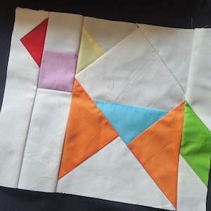 Puede incluir: Un bloque de patchwork con un diseño geométrico. El bloque incorpora colores como rojo, rosa, amarillo, naranja, azul y verde, sobre un fondo blanco. El diseño está compuesto por triángulos y cuadrados.