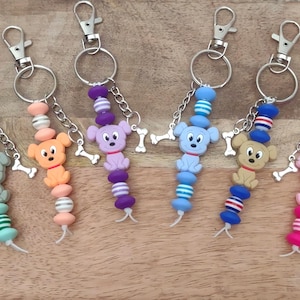 May include: A collection of colorful dog-themed keychains. Each keychain features a cartoon dog charm, a bone charm, and a beaded strand in various colors like green, orange, purple, blue, and pink. The keychains have silver-colored clasps.
