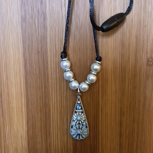 May include: A necklace featuring a black cord with a black barrel clasp, six white faux pearls, and a silver-toned teardrop pendant. The pendant contains a filigree design with light blue and white floral accents and small embedded blue crystals.