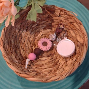May include: A pink and brown beaded keychain with a daisy charm and a retractable badge holder. The keychain is displayed in a woven basket with a pink flower and green leaves in the background. The background is a teal plate.