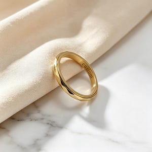 May include: A gold-toned ring with a faceted, geometric outer surface and a smooth inner band. The interior of the ring features the word custom engraved in a clear, lowercase serif font, showcasing a polished metallic finish throughout.