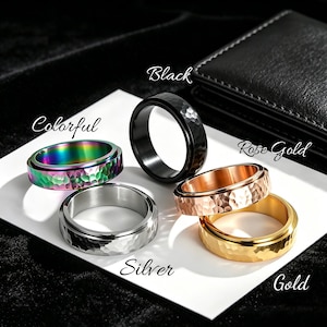 Könnte beinhalten: Fünf Spinner-Ringe mit gehämmertem Finish in verschiedenen Farben: Schwarz, Silber, Roségold, Gold und ein farbenfrohes, schillerndes Finish. Die Ringe werden auf einer weißen Oberfläche mit den Farbbezeichnungen in Schreibschrift präsentiert.
