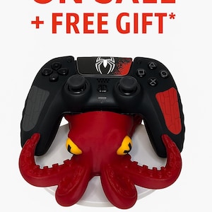 Puede incluir: Mando de videojuegos negro y rojo con el logotipo de Spider-Man, colocado sobre un soporte rojo con forma de pulpo y ojos amarillos. El texto "ON SALE + FREE GIFT*" se muestra arriba.