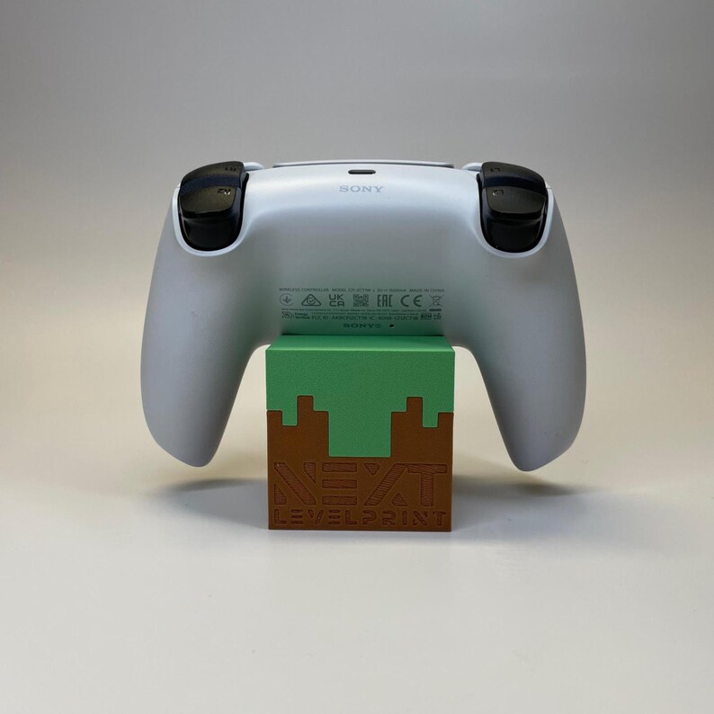 Peut inclure: Une manette blanche de PlayStation 5 repose sur un support en forme de bloc vert et marron. Le support porte l'inscription "NEXT LEVEL PRINT". La manette a des boutons noirs et le logo Sony.