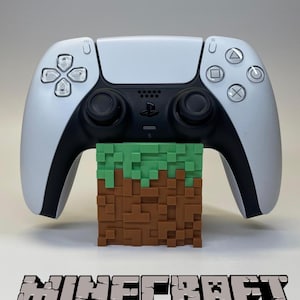 Puede incluir: Un mando blanco y negro de PlayStation 5 descansa sobre un bloque pixelado marrón y verde, que recuerda a un paisaje de Minecraft. La palabra "Minecraft" se muestra en una fuente pixelada 3D debajo.