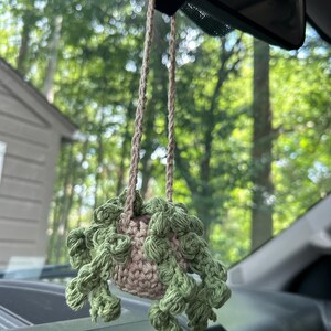 Puede incluir: Un adorno de coche hecho a mano, de ganchillo, con una pequeña maceta marrón claro con detalles de planta verde y frondosa. El adorno cuelga de un cordón trenzado de color beige, suspendido del espejo retrovisor de un coche.