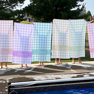 May include: A collection of personalized gingham beach towels in various pastel colors, including pink, purple, blue, and green. Each towel features a name in a matching color on a solid band.