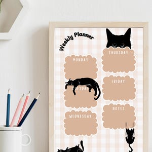 Peut inclure: Un planificateur hebdomadaire encadré avec un cadre en bois clair. Le planificateur présente un fond à carreaux beiges et blancs et des illustrations de chats noirs. Les mots "Weekly Planner" sont en haut. Chaque jour de la semaine est étiqueté.