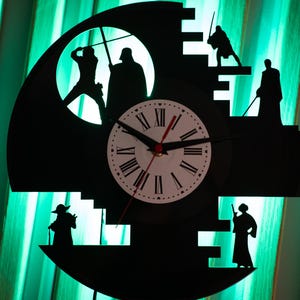 May include: Black wall clock featuring silhouettes of characters from a science fiction movie. The clock face has Roman numerals and red hands. The clock is set against a green-lit background.