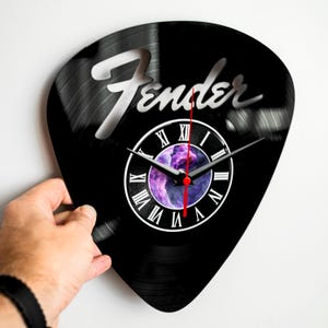 Può includere: Un orologio da parete nero a forma di plettro per chitarra, con il logo Fender. Il quadrante presenta numeri romani e un design galattico viola e blu. Le lancette sono nere con una lancetta dei secondi rossa.
