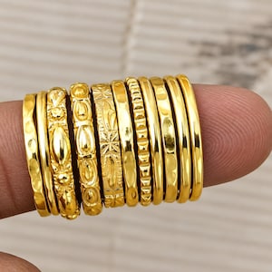 May include: A collection of ten gold-toned rings featuring various textures and patterns, including hammered, floral, geometric, and smooth finishes. The rings are arranged side-by-side, showcasing a variety of decorative metalwork designs.