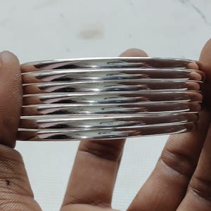 May include: A set of seven stacked silver-toned bangles with a smooth, polished, and reflective metallic surface. The circular bracelets are uniform in width and thickness, featuring a simple, clean design that creates a layered appearance.