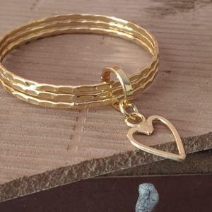 Op de afbeelding: Een set van drie goudkleurige stapelringen met een gehamerde textuur. Een enkele goudkleurige hartvormige bedel is aan de ringen bevestigd met een kleine ronde verbindingsring. De ringen zijn dun en rond met een gepolijste, glanzende afwerking.